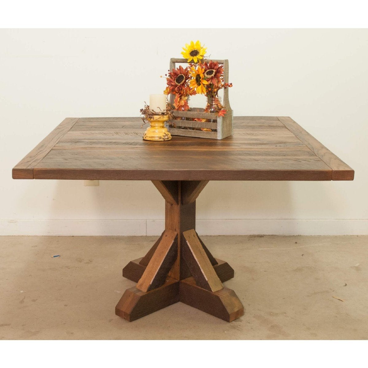 Wood farmhouse table with pedestal base, sunflowers, blooms, candles; plain white wall behind.