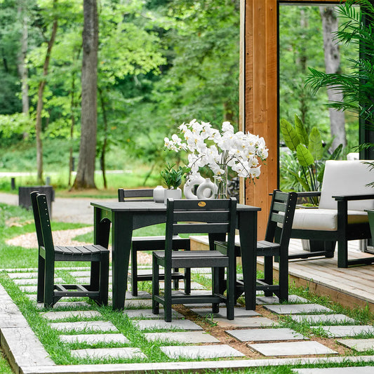 Black outdoor dining set with vase, chairs on patio, greenery and trees in the background.