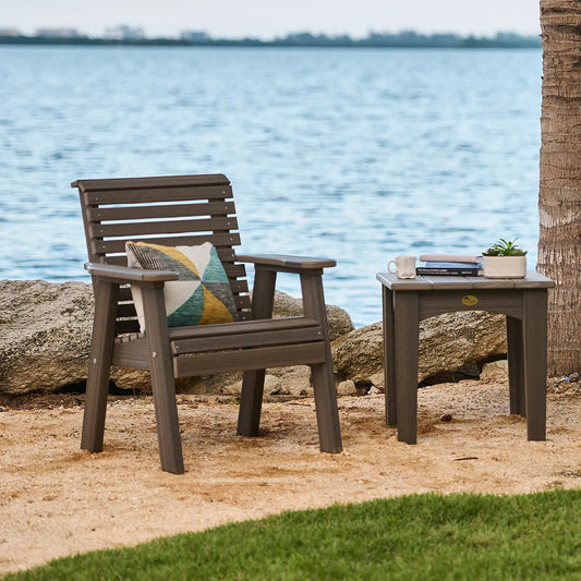 Bench and small table with decor on sand by water, rocks, and grass—cozy outdoor setup.