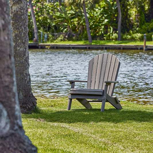 Folding Adirondack chair on grass by a calm river, shaded by trees; lush greenery in background.