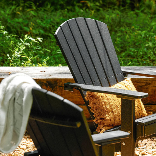 Black Adirondack chair with yellow pillow outdoors; white blanket and greenery in the background.