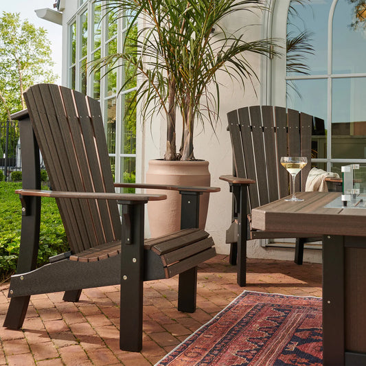 Two Adirondack chairs and a small drink table sit on a brick patio by lush greenery and windows.