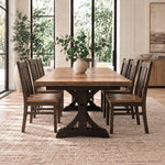Wooden dining table with six chairs, vase centerpiece, on beige rug near windows and a plant.