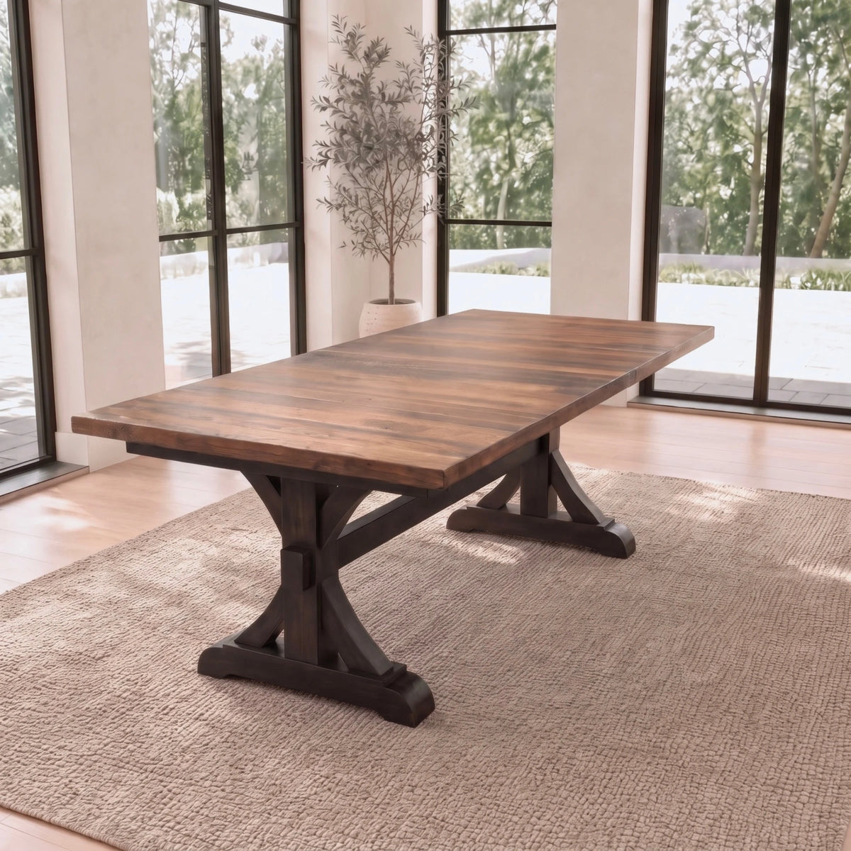 Solid wood dining table with dark base on beige rug in bright room, tall windows, potted plant.