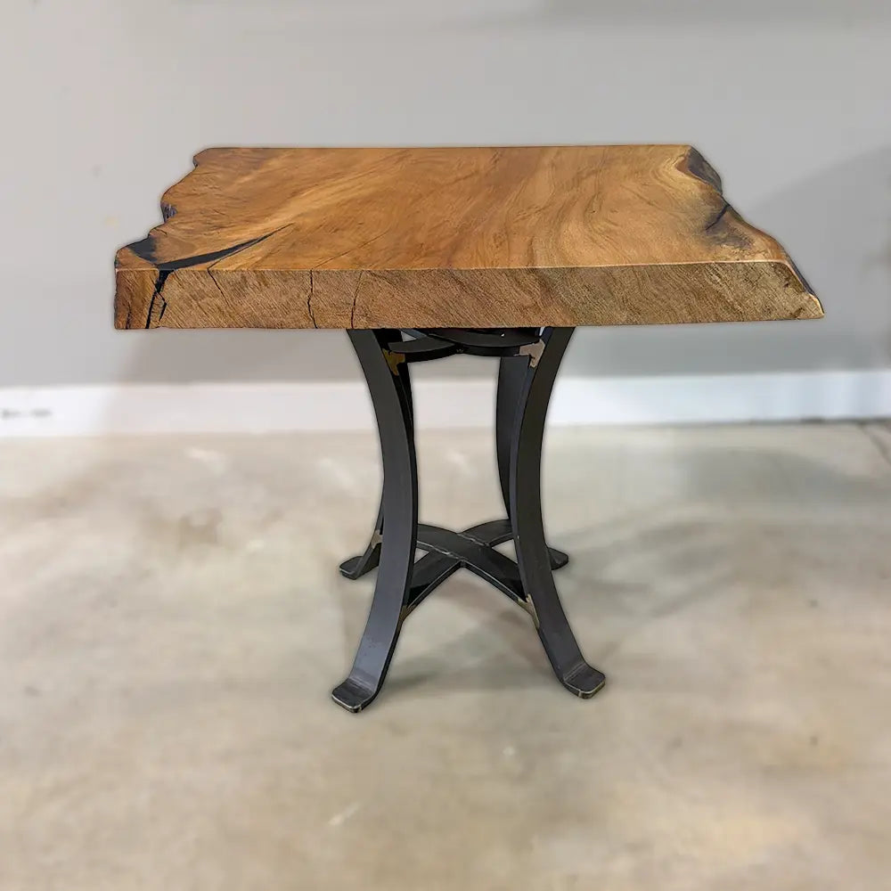 The 25" Live Edge Sycamore End Table features a thick, live edge sycamore wood slab top with natural cracks and a smooth polyurethane finish, supported by a black steel base with gracefully curved legs and set on a smooth concrete floor.