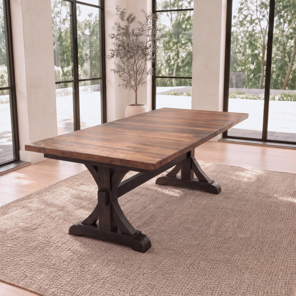 Solid wood dining table on woven rug in bright room with large windows and potted plant.
