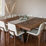 Square oak dining table with eight white chairs, glass jars, and pampas grass by the window.