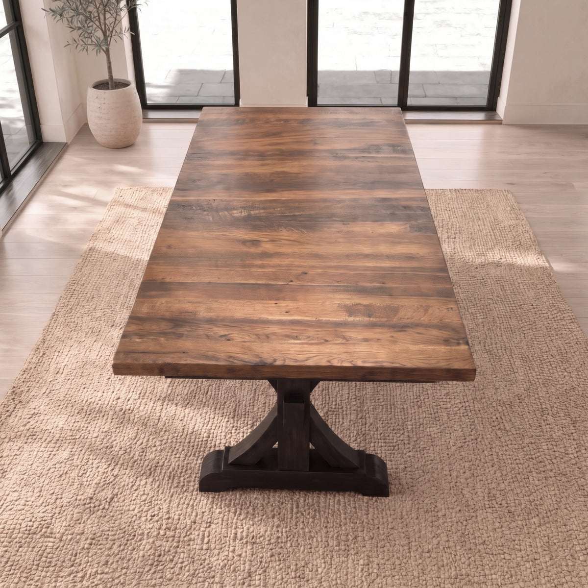 Wood dining table with trestle base on beige rug in bright room with windows and potted plant.