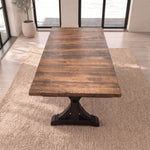 Wood dining table with trestle base on beige rug in bright room with windows and potted plant.