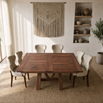 Square dining table with six chairs on a rug, in a bright room with shelves and a macramé hanging.