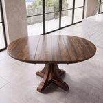 Round reclaimed wood dining table with pedestal base by windows, tiled floor, balcony view.