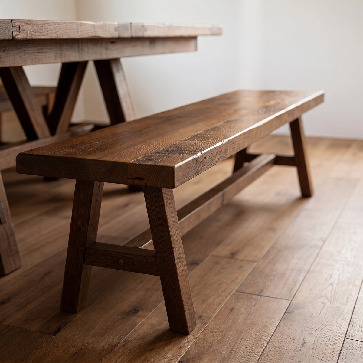 Rustic barnwood bench with rectangular top and splayed legs on a wood floor by a table.