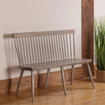 Colby Windsor Bench with spindle back, angled legs; by potted plant on wood floor, driftwood finish.