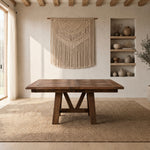 Oak dining table on woven rug in bright room with macramé art, shelves with pottery, wood beams.