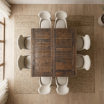 Top-down view of a square oak dining table with 8 cream chairs on a beige rug in a bright room.