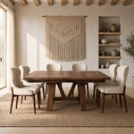 Rustic dining room with a wood table, beige chairs, woven rug, macramé art, and natural light.