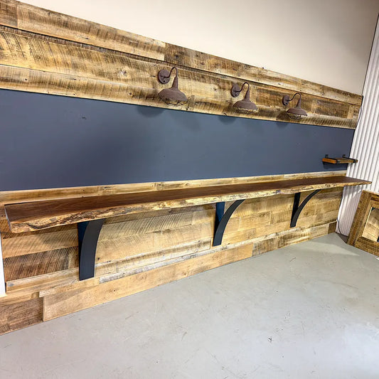 Live edge walnut bar top with black brackets, lit by 3 lamps on rustic wood wall, gray floor.