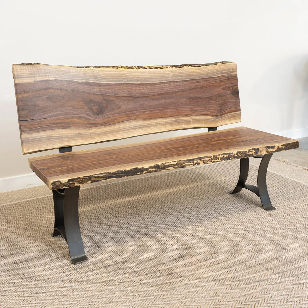 Live edge wood bench with back and black metal legs on beige rug against a white wall.