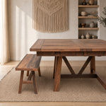 Square oak dining table and bench on rug; shelves with pottery, plant, macrame, sunlight filters in.