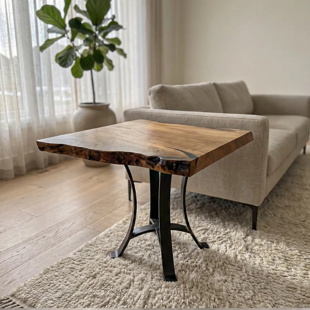 The 25" Live Edge Sycamore End Table, featuring a smooth polyurethane-finished sycamore wood top with distinctive live edges and black steel base legs, is placed beside a beige sofa on a light rug in a bright living room adorned with sheer curtains and a potted plant, showcasing the wood's natural beauty.