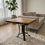 The 25" Live Edge Sycamore End Table, featuring a smooth polyurethane-finished sycamore wood top with distinctive live edges and black steel base legs, is placed beside a beige sofa on a light rug in a bright living room adorned with sheer curtains and a potted plant, showcasing the wood's natural beauty.