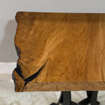 A close-up of the 25" Live Edge Sycamore End Table highlights its natural grain patterns and distinctive blackened cracks on the left. The table is supported by a sleek Natural Steel Base and rests on a light-colored floor, showcasing its unique charm.