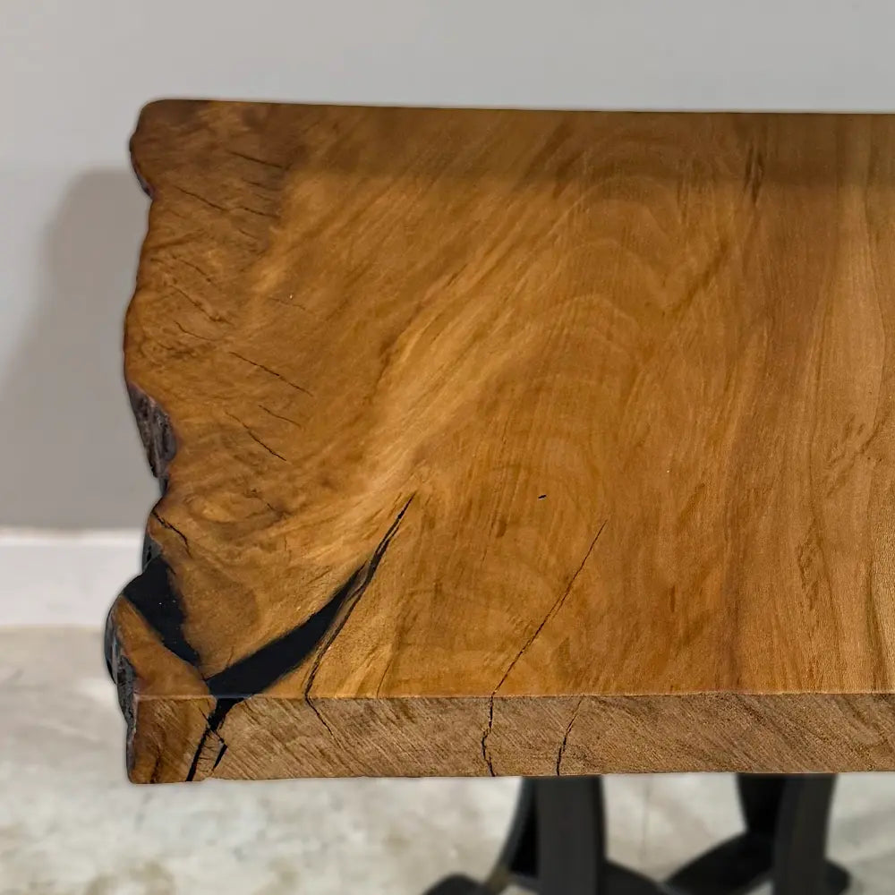 A close-up of the 25" Live Edge Sycamore End Table highlights its natural grain patterns and distinctive blackened cracks on the left. The table is supported by a sleek Natural Steel Base and rests on a light-colored floor, showcasing its unique charm.