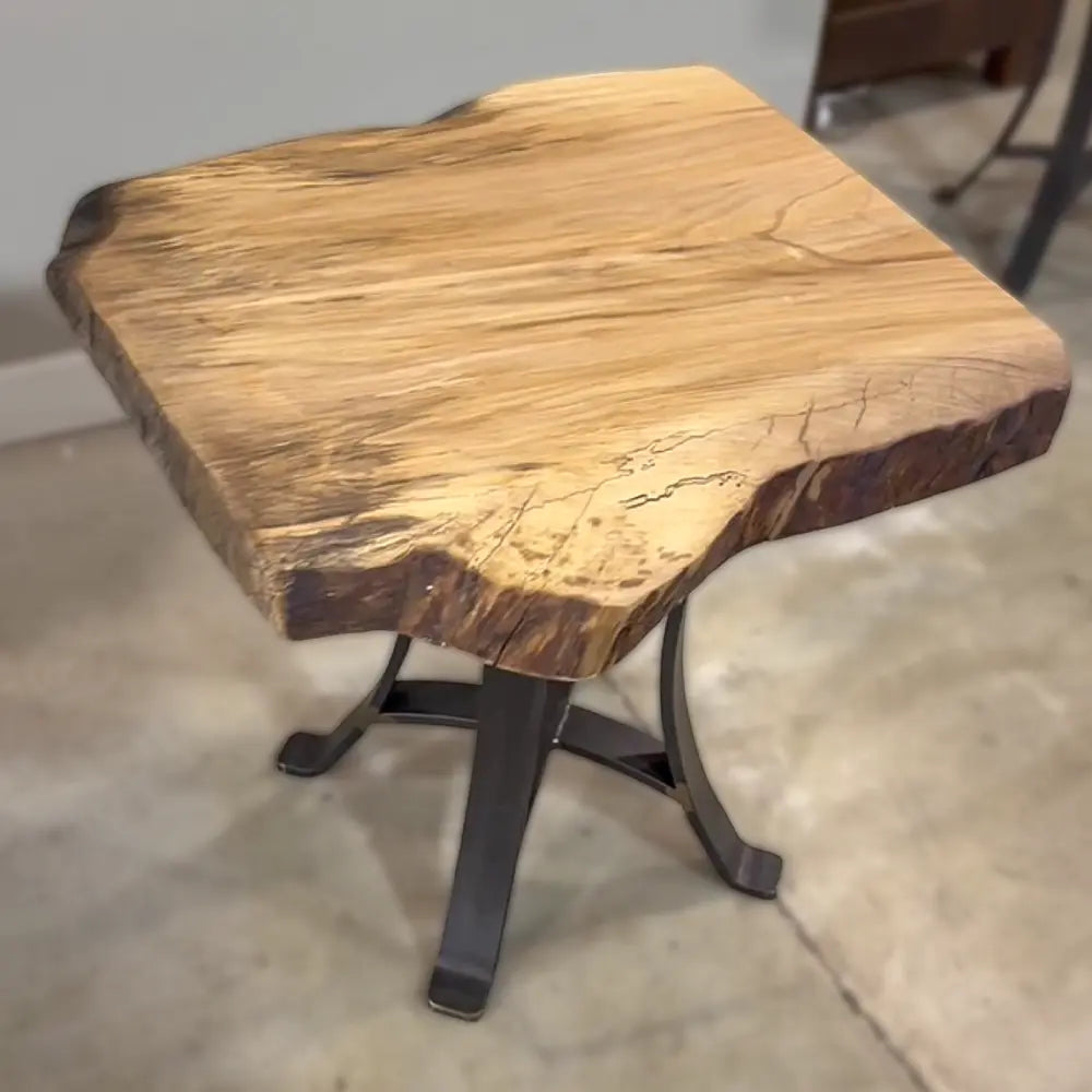 Live Edge Sycamore End Table with black steel base on concrete floor by a light wall.