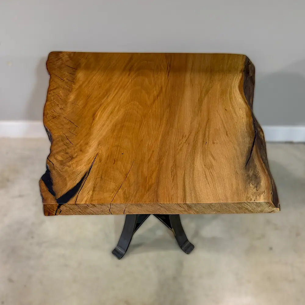 The 25" Live Edge Sycamore End Table showcases a beautifully crafted live edge sycamore wood top with a smooth polyurethane finish, supported by bold black metal legs. It is displayed on a light-colored floor against a plain gray wall.