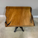 The 25" Live Edge Sycamore End Table showcases a beautifully crafted live edge sycamore wood top with a smooth polyurethane finish, supported by bold black metal legs. It is displayed on a light-colored floor against a plain gray wall.